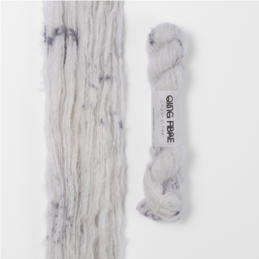 Platinum Melted Baby Suri 25g: white with grey markings yarn, shown loose beside a labelled hank.