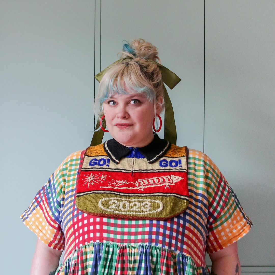 Woman with blonde hair and blue streak, wearing colorful checkered dress and bib reading 2023