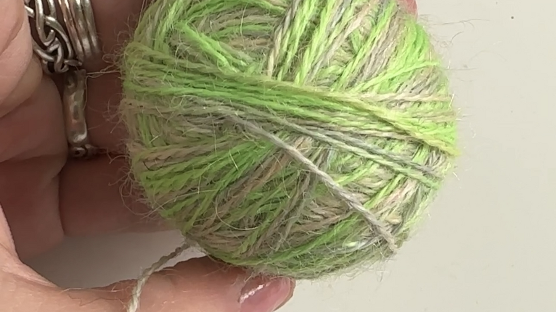 Hand holding a ball of green and beige yarn against a light background