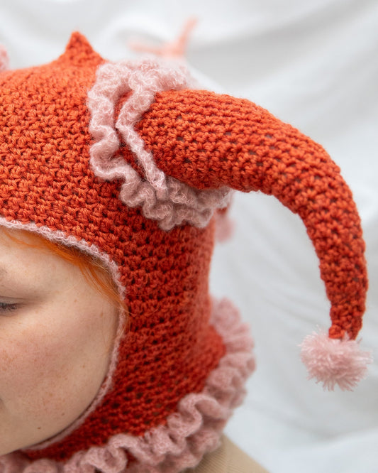 Close-up of person wearing a red and pink crochet jester hat with ruffles and pom-poms