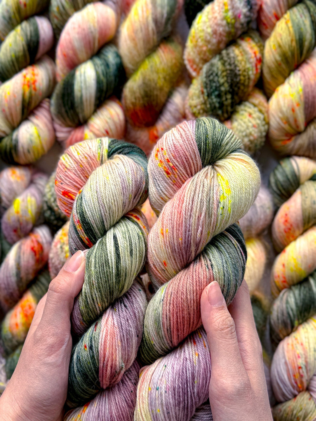 Hands holding two skeins of multicolored hand-dyed yarn with more skeins in the background