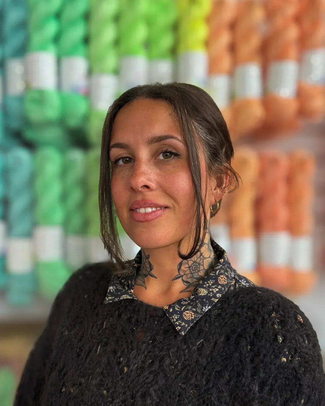 Woman with neck tattoo in black sweater, standing in front of colorful yarn skeins display