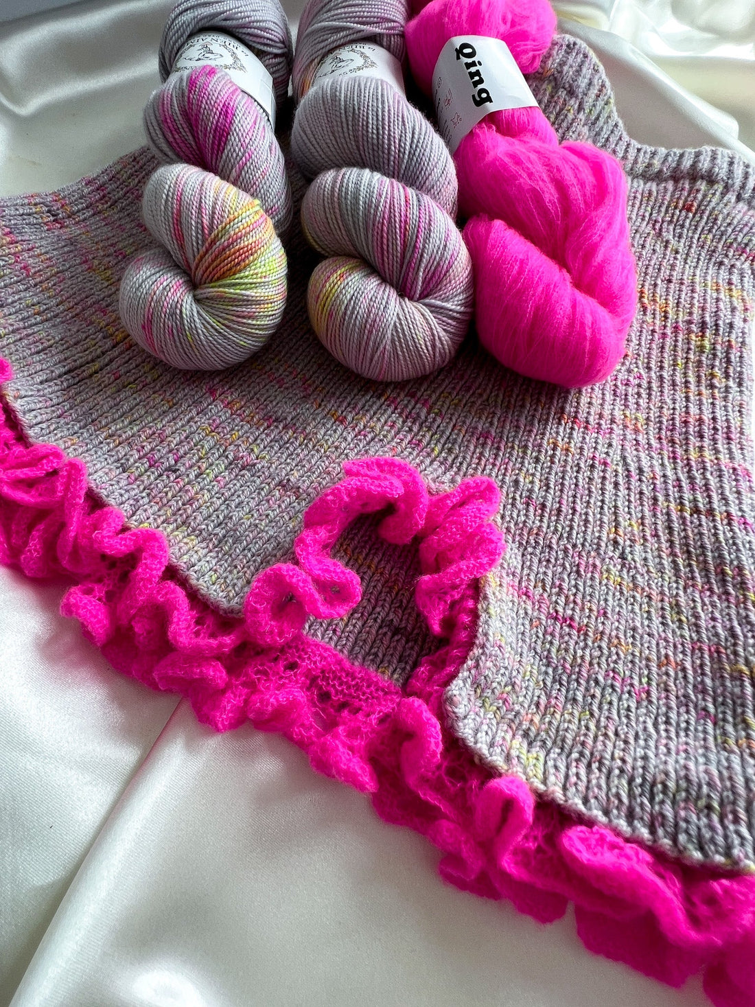 Hand-knitted gray sweater with pink ruffle trim and skeins of pink and gray yarn on satin fabric