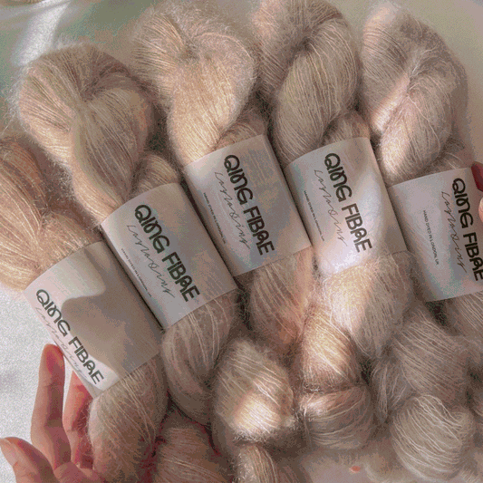 Hand holding skeins of pale beige mohair yarn labeled QING FIBRE luxuriously soft