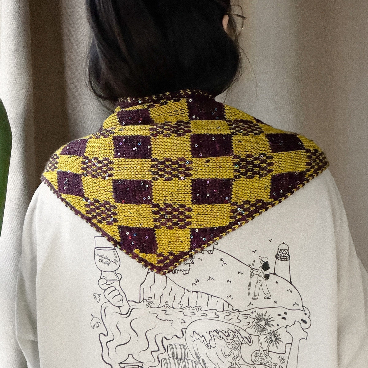 Harlequin Gambit triangle scarf worn over the shoulders, showing burgundy and yellow checks with sequins.