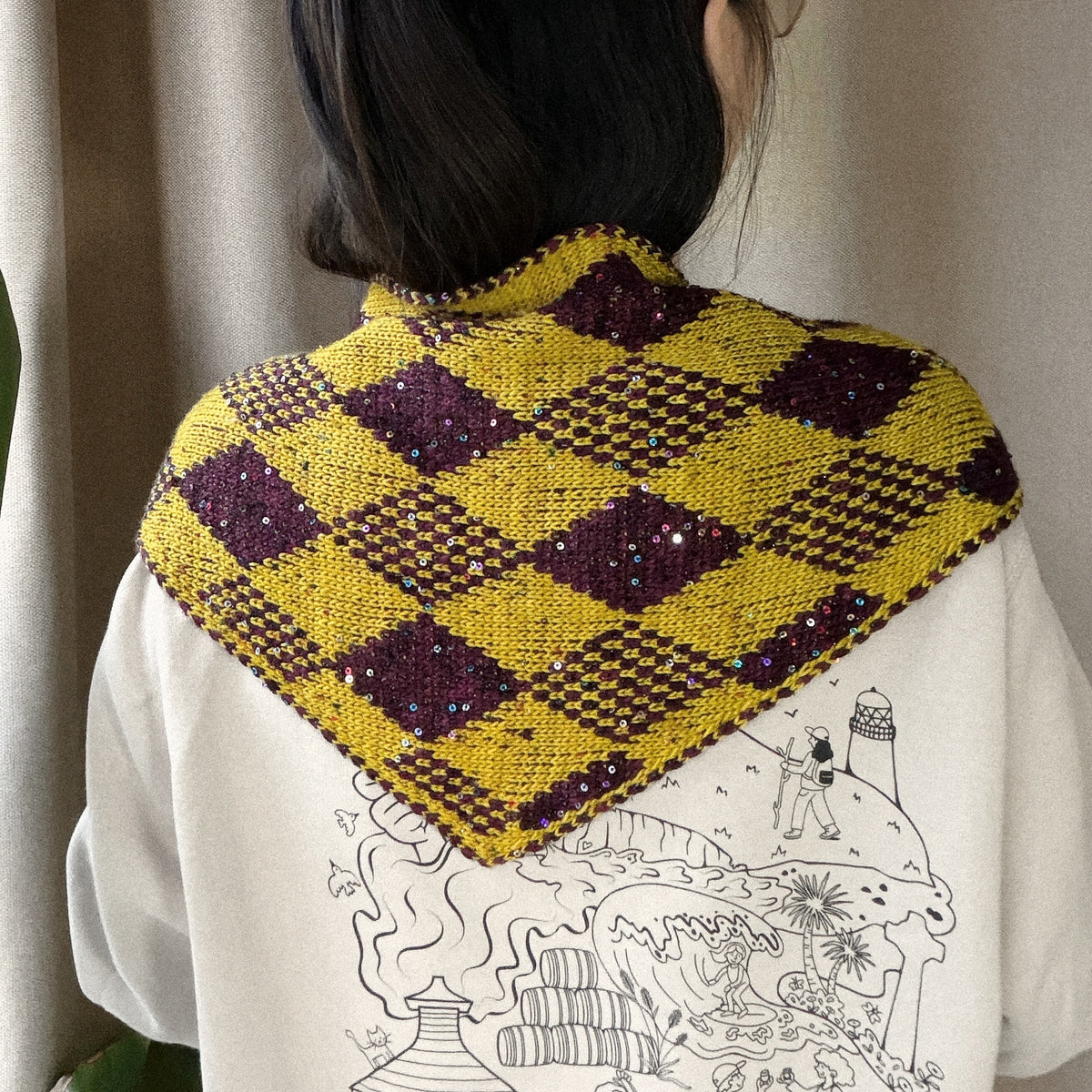Harlequin Gambit triangle scarf worn over the shoulders, showing burgundy and yellow diamond motifs with sequins.